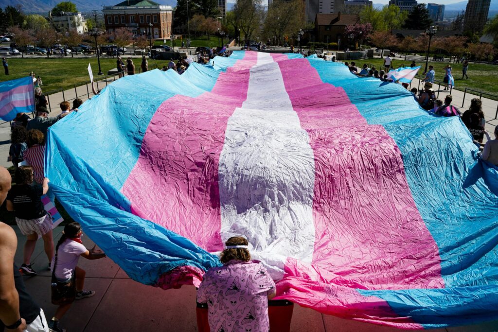 People holding a large transgender pride flag outdoors, representing community support for HRT in Texas