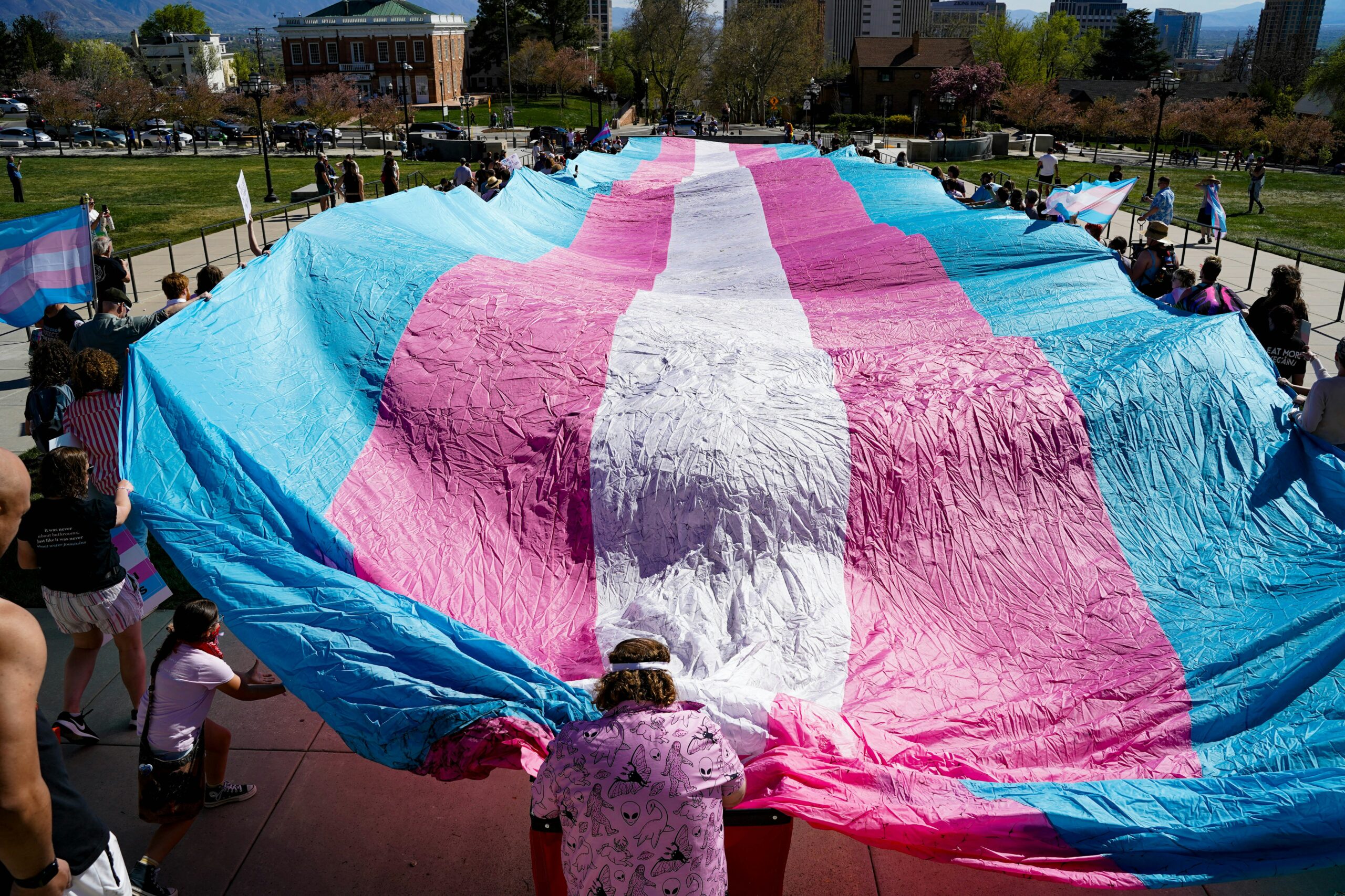 People holding a large transgender pride flag outdoors, representing community support for HRT in Texas