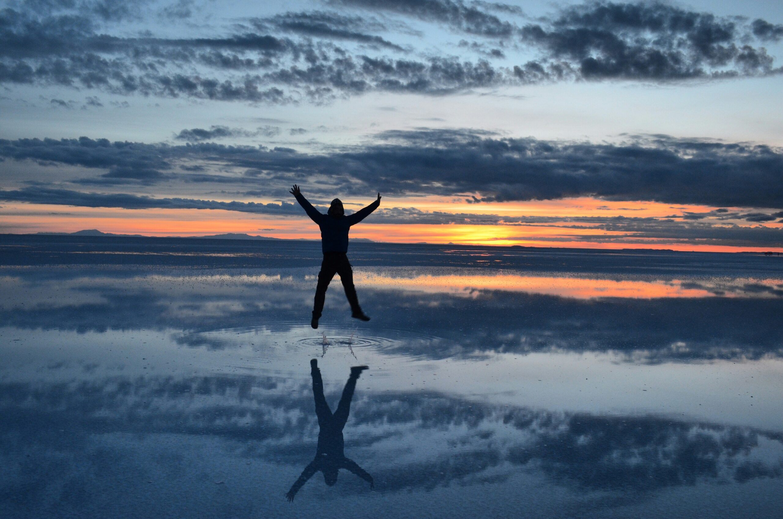 A person silhouetted against a sunset sky, standing with both arms raised fully overhead, representing the full range of motion recovery can restore